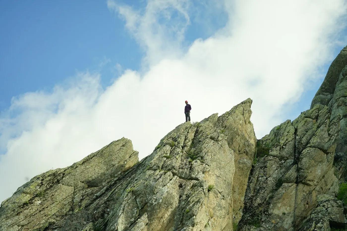 A lone hiker stands atop a rocky mountain peak under a bright blue sky.