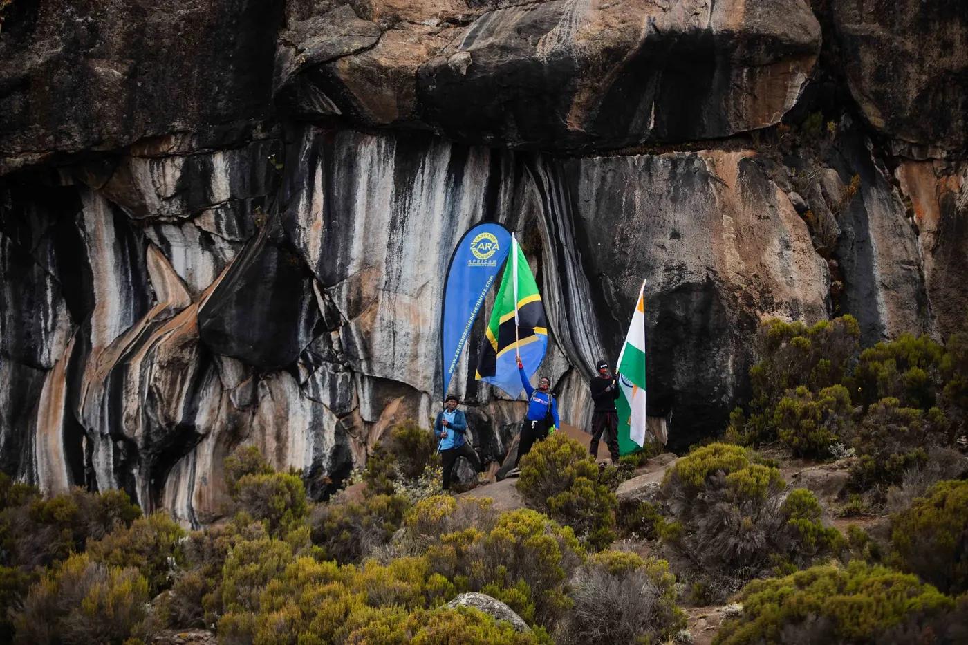 Hikers holding Tanzanian and other flags at a cave entrance on Mount Kilimanjaro.