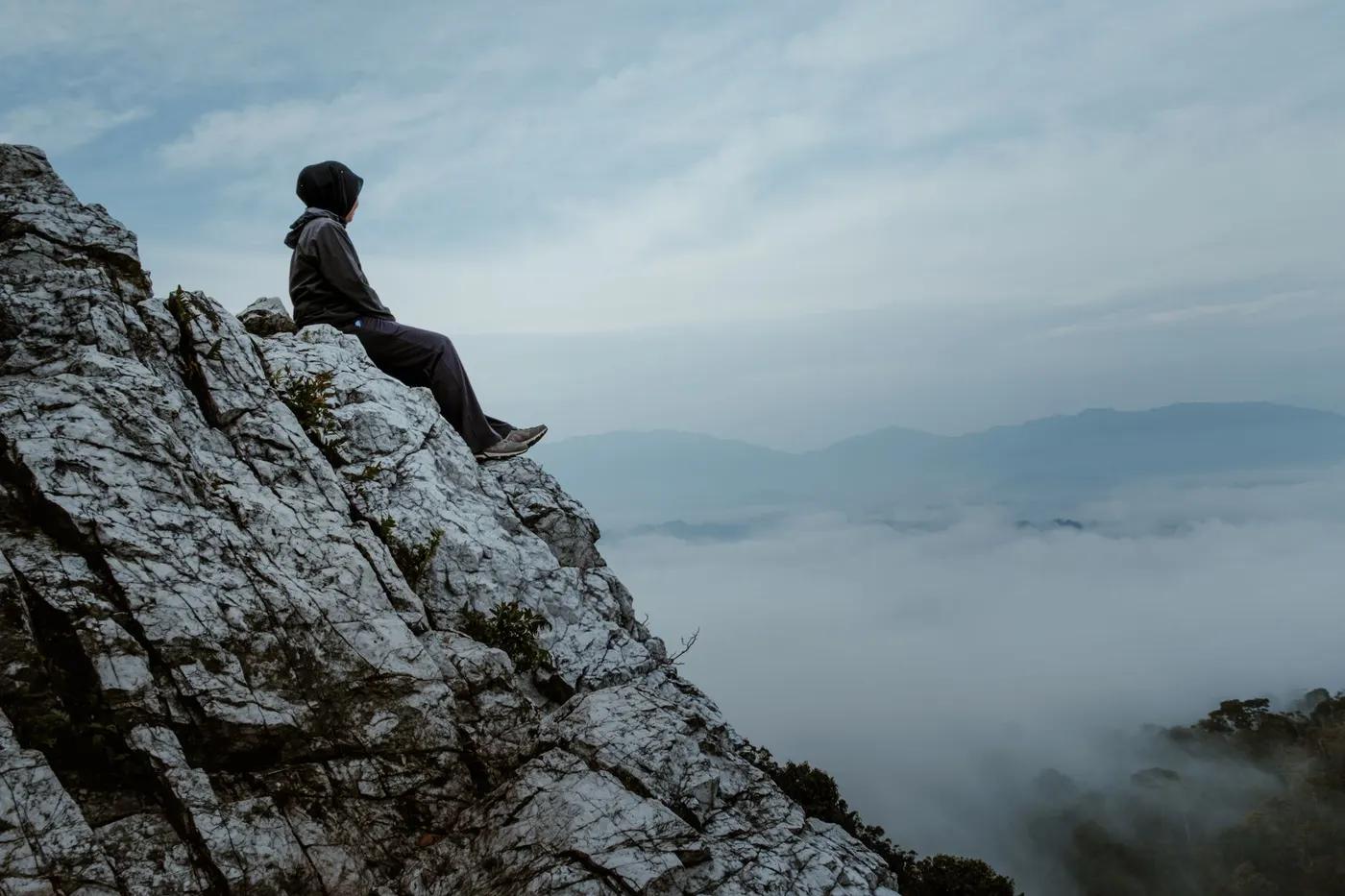 A lone hiker sits on a rocky mountain peak in Kuala Klawang amidst a scenic misty landscape.