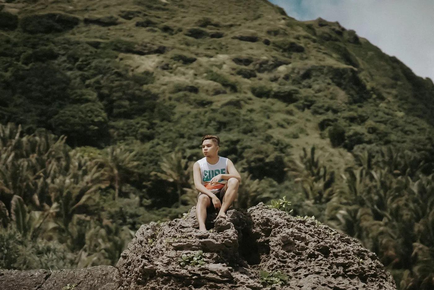 Adult man sitting on a rock with lush mountain backdrop in Basco, Cagayan, Philippines.