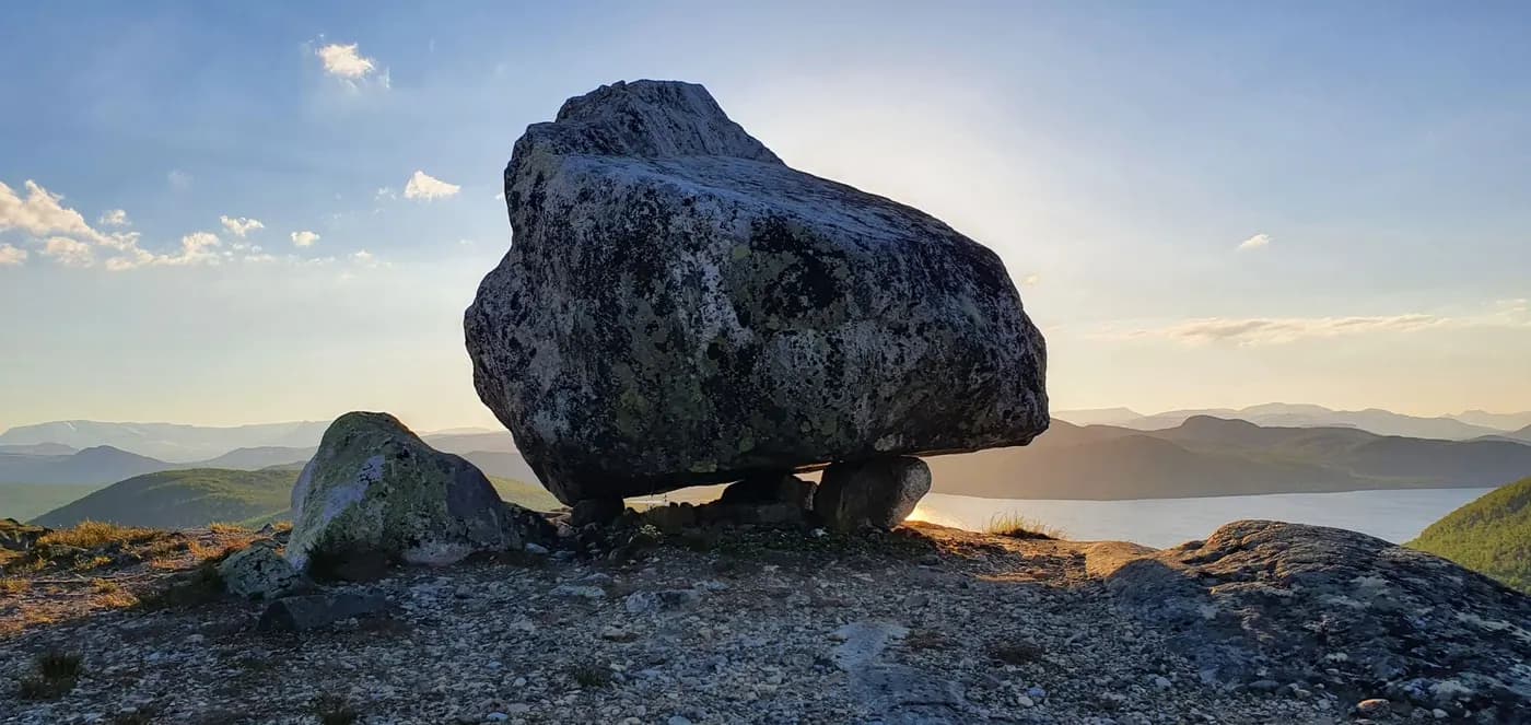A large balancing rock at sunset with a scenic view of mountains and a lake.