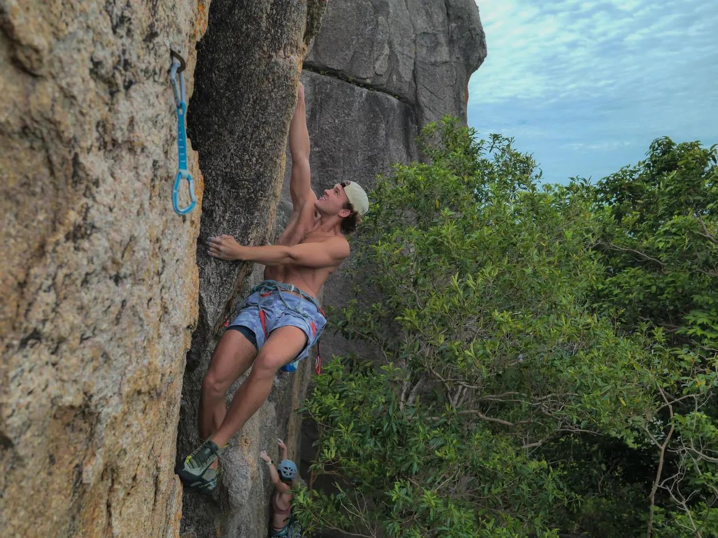 A man climbs a steep rock face surrounded by greenery in Ko Tao, Thailand.