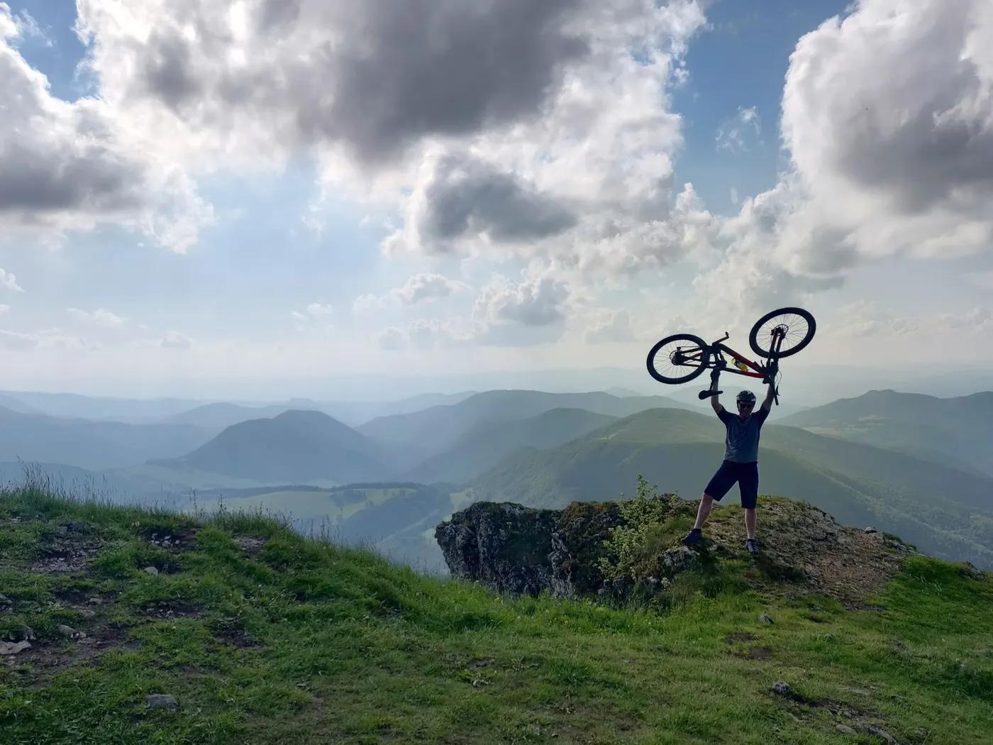 A mountain biker triumphantly lifts their bike atop a scenic hill with expansive views.