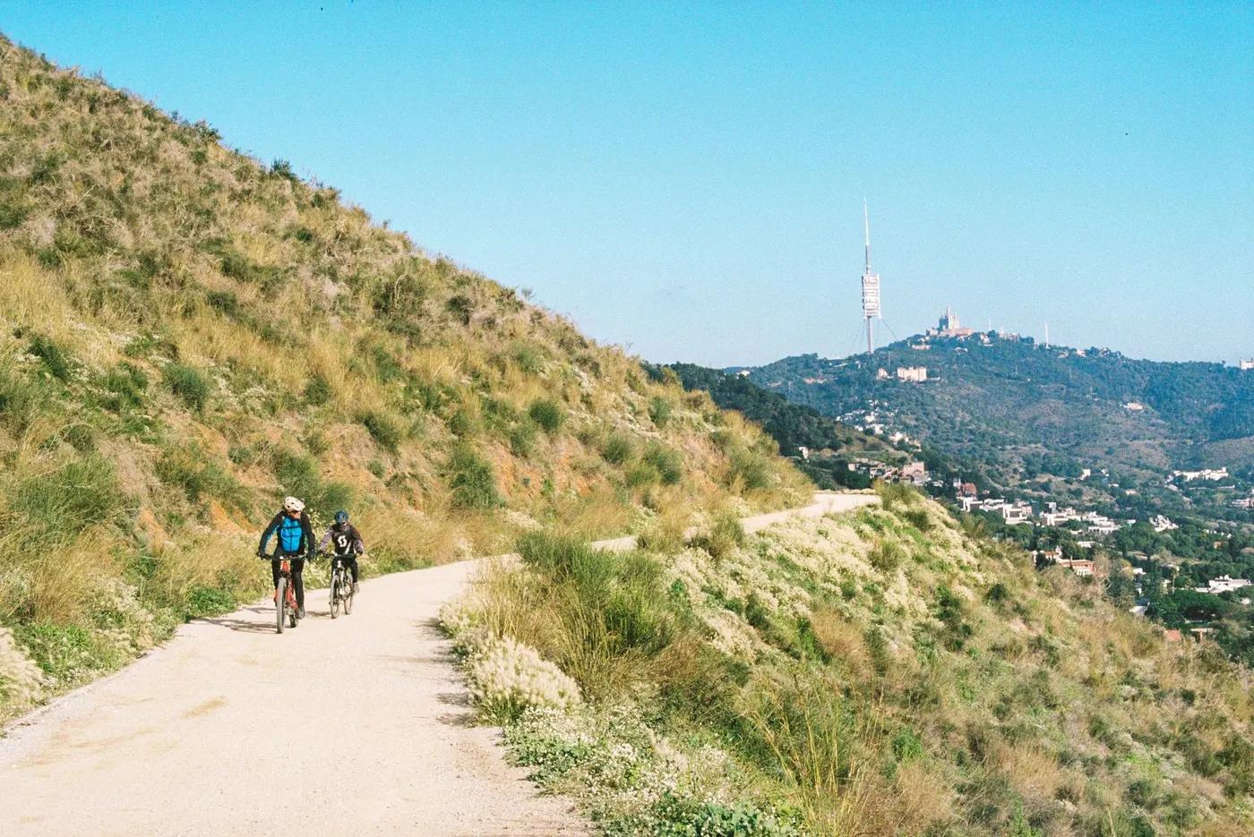 Cyclists ride along a mountain trail with distant views of a prominent TV tower.