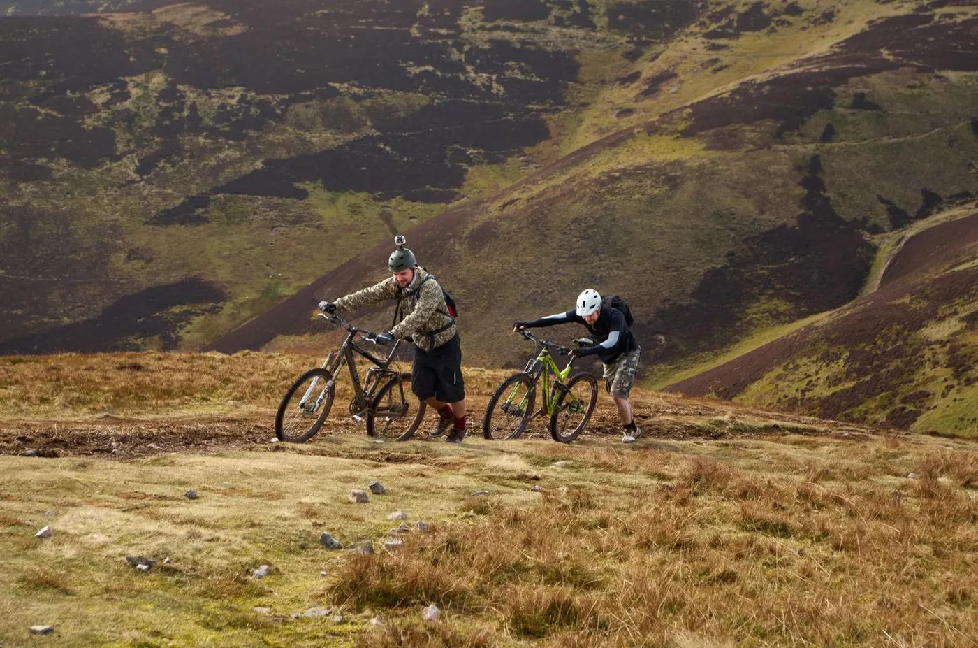 Two men pushing mountain bikes uphill in Scotland's scenic Pentland Hills.