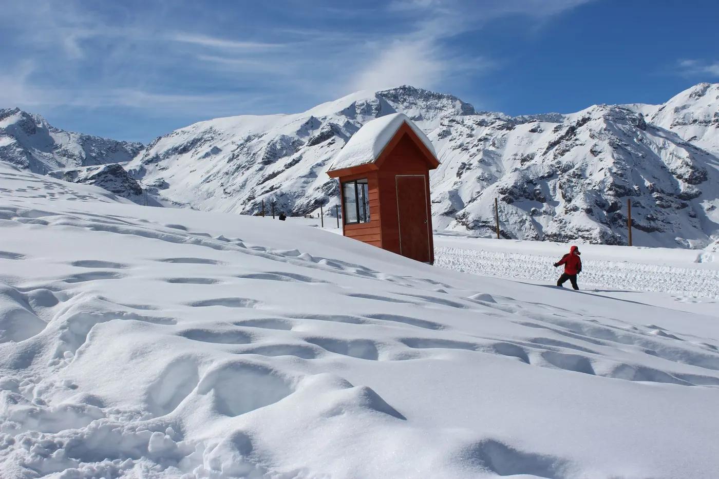 A skier clad in winter gear traverses a snowy mountain slope beside a small wooden cabin.