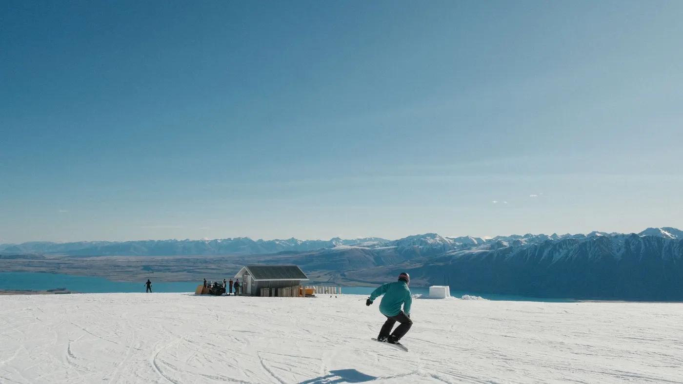 Snowboarder descends a snowy slope with a stunning mountain and lake view on a clear winter day.