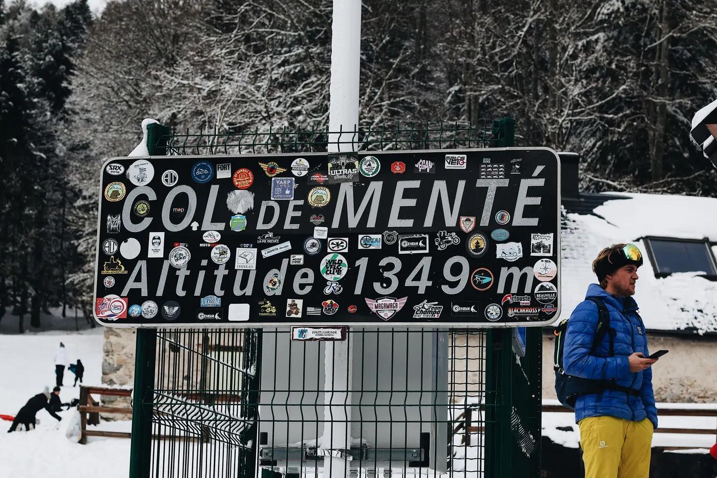 Sign at Col de Menté mountain pass surrounded by snowy trees and winter scenery.