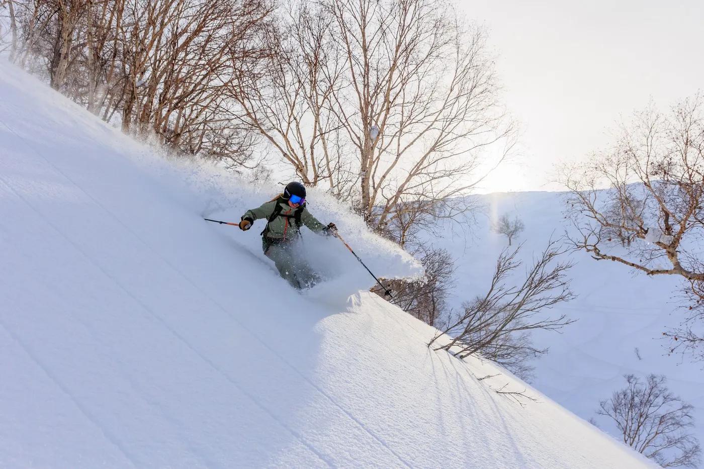 A skier swiftly descends a snow-covered slope surrounded by bare trees under a clear sky.