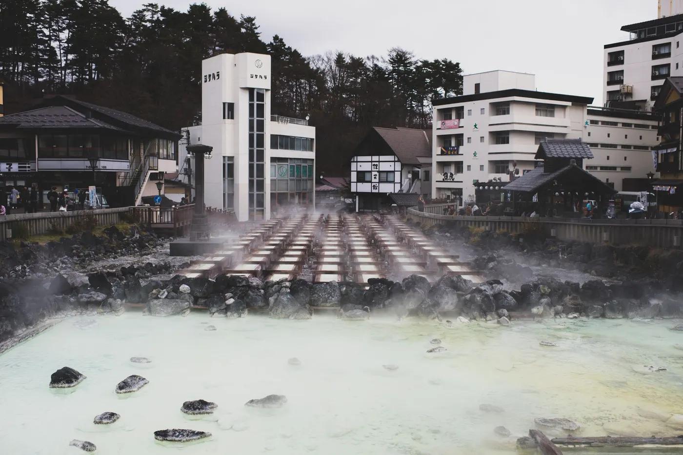 A tranquil hot spring town with steam rising from mineral-rich waters, surrounded by traditional architecture.
