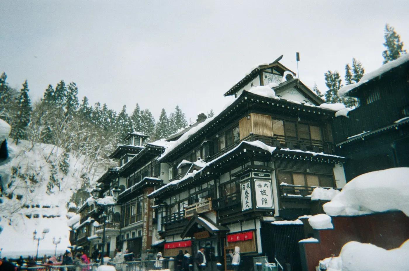 A picturesque view of traditional Japanese architecture in a snowy onsen town, set against a backdrop of snow-covered t…