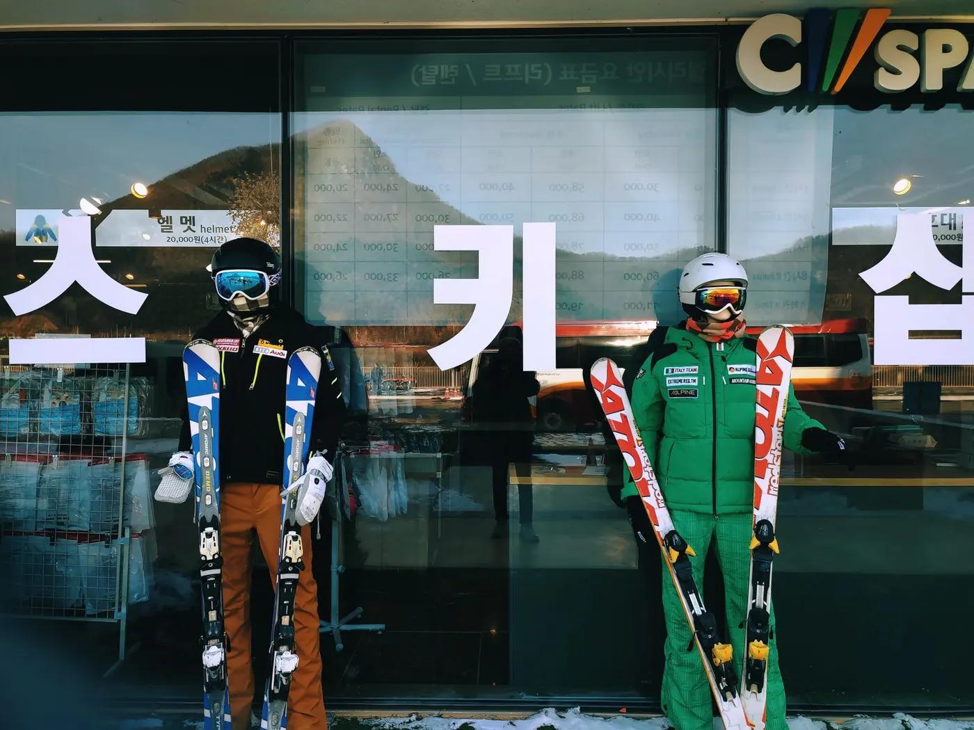 Skiers in winter gear holding skis in front of a ski shop with reflections and snowy backdrop.