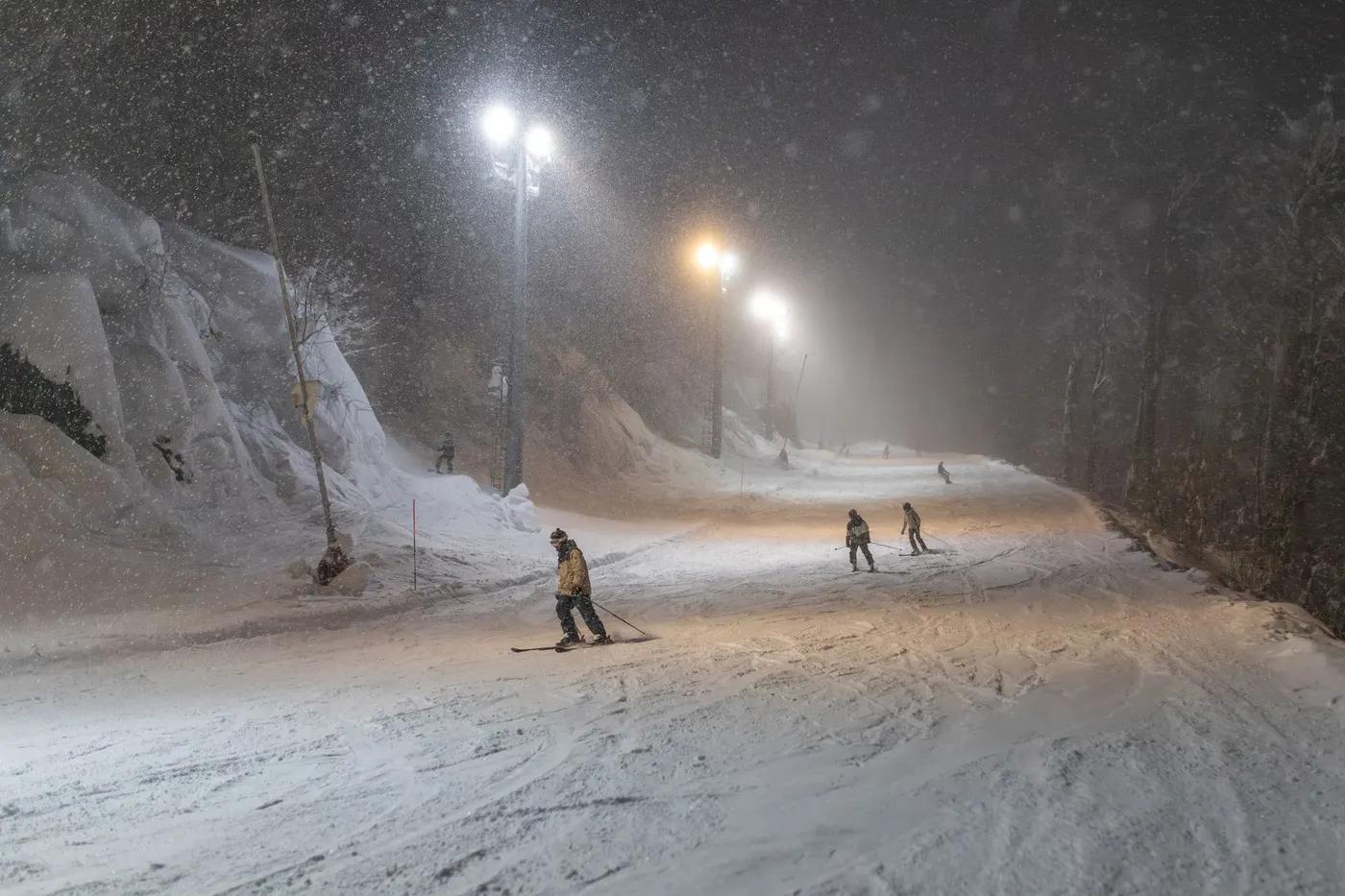 Skiers enjoy a snow-filled night on a well-lit mountain slope.
