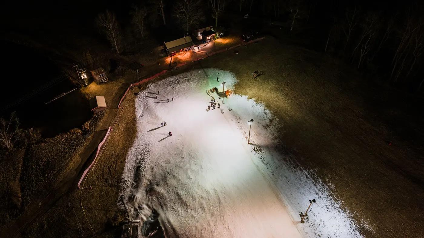 Aerial shot of a night-time ski slope with artificial snow, illuminated by lights and small crowd gathered.