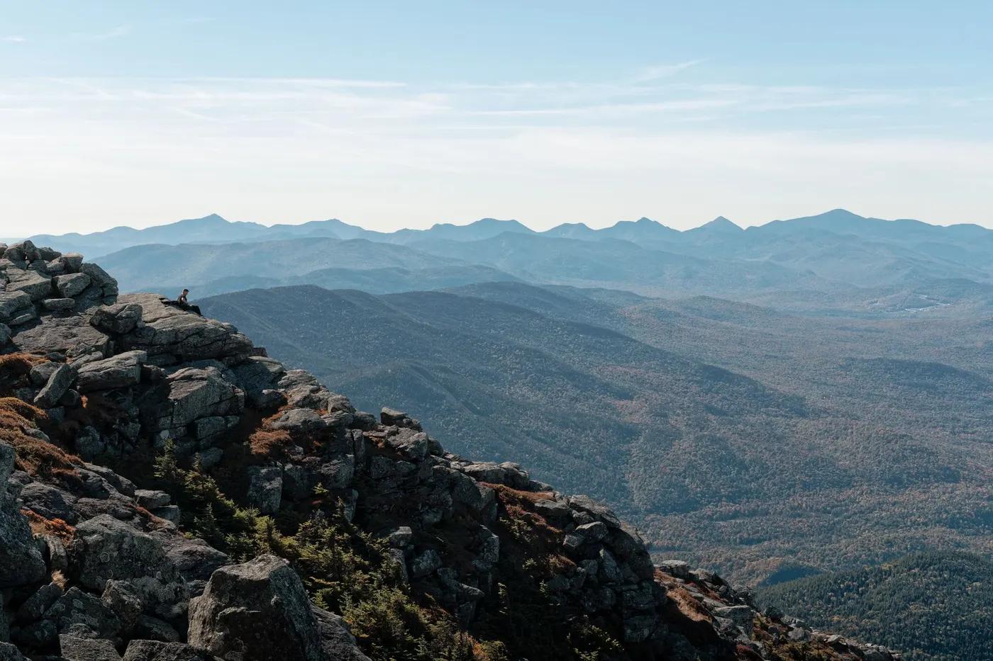 A lone hiker admires the vast landscape of the Adirondack Mountains under a clear blue sky.