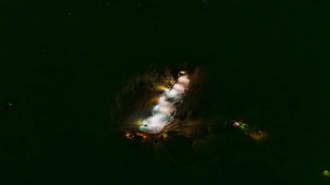 Aerial shot of a brightly lit ski slope at night, surrounded by dense forest.